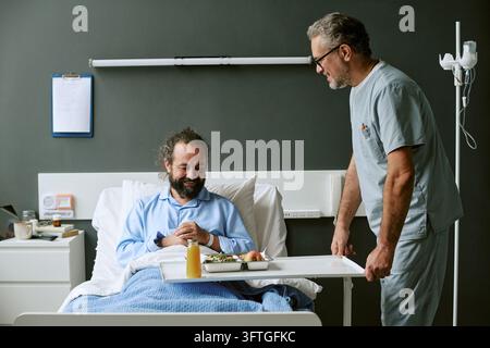 Infermiere maschio di mezza età che serve cibo a un paziente in un reparto ospedaliero Foto Stock