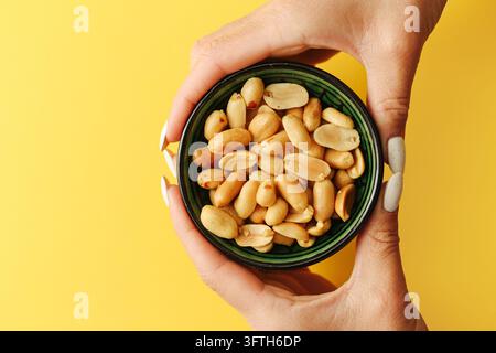 Mani che tengono una ciotola verde di arachidi arrostite su sfondo giallo. Spuntino sano con noci croccanti, proteine vegetali e cibo nutriente per tutti i giorni. Foto Stock