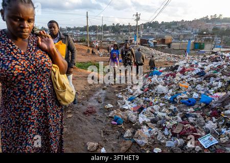 Persone che camminano lungo un sentiero fangoso vicino a un cumulo di rifiuti con una donna in abito a fiori in primo piano nell'area di Kibera, Contea di Nairobi, Nairobi, Kenya Foto Stock