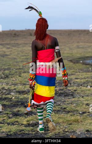 Vista posteriore di un uomo della tribù Rendille che cammina su un pascolo roccioso con abiti colorati e calzini a righe, Contea di Marsabit, Loiyangalani, Kenya Foto Stock
