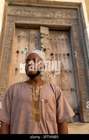 Uomo barbuto con una kofia bianca ricamata e una camicia marrone ricamata davanti a una vecchia porta di legno intagliata di Zanzibar, Stone Town, Tanzania Foto Stock
