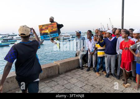 Uomo che salta a mezz'aria tenendo un dipinto con la scritta "Benvenuti a Zanzibar" mentre una folla di ragazzi e uomini guarda al crepuscolo, Stone Town, Tanzania Foto Stock