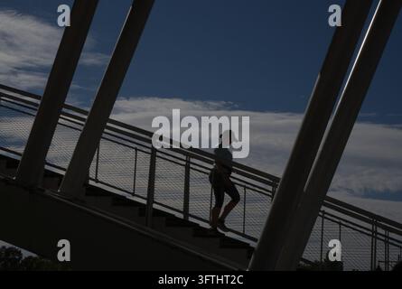 Silhouette di una persona che sale le scale di un moderno ponte pedonale contro un cielo azzurro con soffici nuvole bianche in Francia Foto Stock