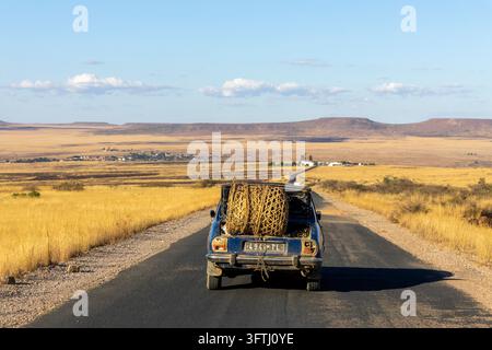 Una vecchia auto con un grande fascio di paglia intrecciata legato al bagagliaio aperto percorre una strada rurale attraverso la savana, Ihorombe, Isalo, Madagascar Foto Stock