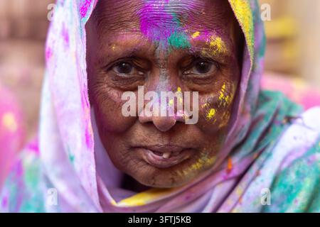Anziana donna con rughe profonde e gulal viola e giallo spalmato sul viso guarda nell'obiettivo, Uttar Pradesh, Vrindavan, India Foto Stock
