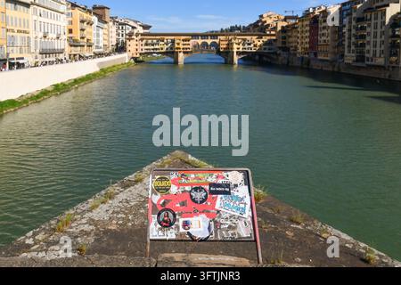 Vista panoramica di Ponte Vecchio sul fiume Arno da un pilone di pietra di Ponte Santa Trinita con un cartello ricoperto di adesivi a Firenze, Toscana, Italia Foto Stock
