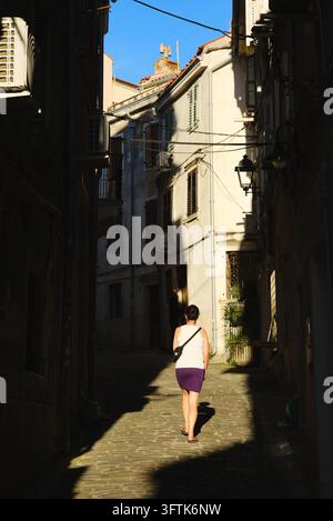Donna che sale per un vicolo stretto e soleggiato lastricato di ciottoli tra case storiche in pietra nella vecchia città medievale di Piran, in Slovenia. Foto Stock