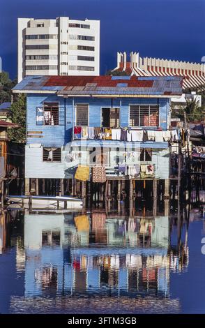 In Malesia, una casa su palafitte con il bucato appeso ad asciugare si riflette nell'acqua calma. sullo sfondo ci sono edifici più moderni. Foto Stock