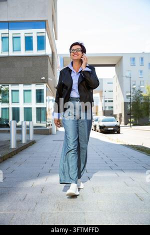 Una donna d'affari matura sorridente parla al telefono mentre tiene un laptop in una strada della città di Helsinki fuori da un ufficio moderno Foto Stock