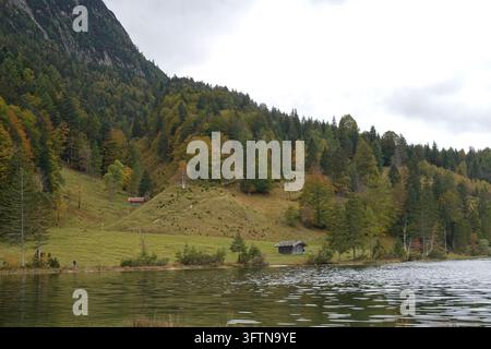 Gli alberi autunnali coprono i pendii delle montagne accanto al lago Ferchensee a Mittenwald, in Baviera, Germania, con una capanna di legno e escursionisti lungo la riva del lago. Foto Stock