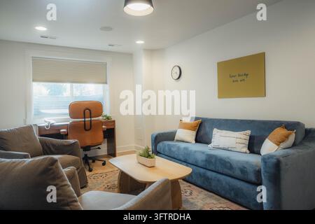 Soggiorno moderno e luminoso con divano in velluto blu, cuscini decorativi a motivi, tavolino da caffè in legno, poltrone grigie e piccola scrivania con sedia da ufficio in pelle Foto Stock