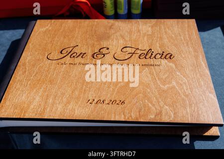 Libro degli ospiti in legno con nomi incisi Jon & Felicia data 12.08.2026 primo piano che mostra la calda venatura del legno e l'incisione dello script sulla copertina appoggiata su uno sfondo scuro Foto Stock