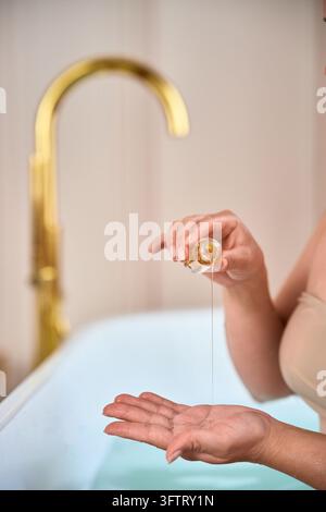 Donna versa olio per il corpo dorato da una piccola bottiglia di vetro in un palmo aperto accanto alla vasca da bagno, preparando una routine di cura idratante con un sottile getto Foto Stock