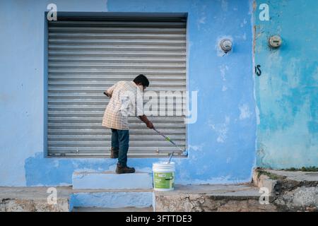 Messico - 11 febbraio 2025: Vista di un uomo che dipinge un muro blu con un rullo davanti a una saracinesca metallica. Foto Stock