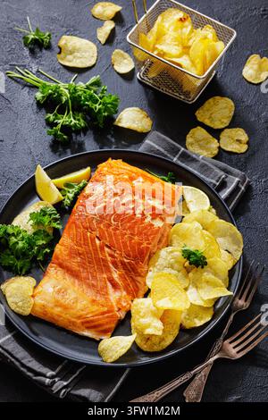 Filetto di salmone affumicato caldo con patatine, fette di limone fresco e prezzemolo verde su un piatto con forchette e cestino per friggere su uno sfondo nero, verticale Foto Stock