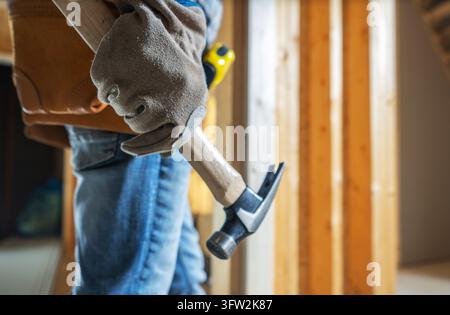 Vista ravvicinata della mano guantata di un falegname che tiene un martello a becco accanto a una cintura degli attrezzi e a una parete in legno a vista. Foto Stock