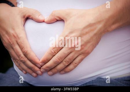 Un marito abbraccia la sua moglie incinta al ventre e forma un cuore con le dita Foto Stock