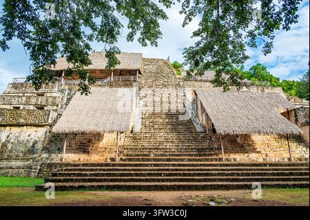 Ampia veduta frontale della piramide principale di Ek Balam nello Yucatan, Messico. L'antica struttura in pietra Maya si innalza in ripidi gradini che conducono a una piattaforma tempio. Tetti di palapa in paglia proteggono alcune parti delle rovine, mentre rami frondosi incorniciano la cima dell'i Foto Stock