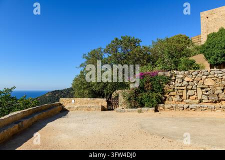 Una terrazza in pietra con muretti bassi si affaccia sul mare a Pigna. Vegetazione mediterranea e architettura rustica del villaggio sotto un cielo blu terso. Foto Stock