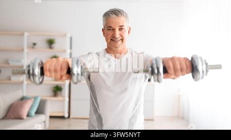 Uomo atletico maturo che solleva i manubri, fa allenamento di forza per le braccia, guarda in camera e sorride, si allena a casa nell'interno del soggiorno, pano Foto Stock