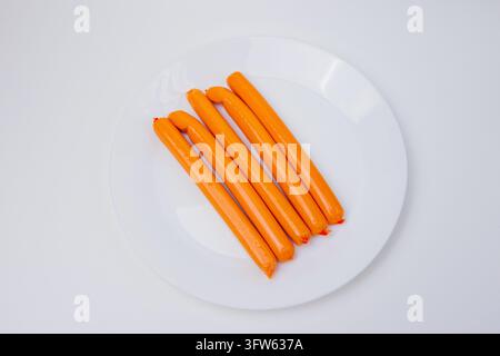 Cinque salsicce arancioni brillanti in involucri lucidi disposte in diagonale su un piatto di ceramica bianco pulito su uno sfondo chiaro. Natura morta alimentare semplice hi Foto Stock
