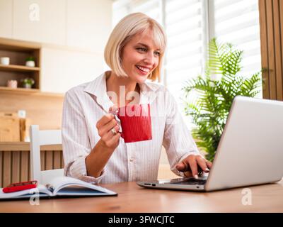 Donna felice che assapora un tranquillo caffè mattutino mentre lavora sul suo laptop. Foto Stock