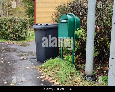 Una cassetta delle poste verde autonoma e un bidone della spazzatura in plastica nera posizionati fianco a fianco all'aperto in una giornata autunnale, posizionati accanto a un percorso in asfalto bagnato Foto Stock