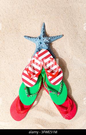 Infradito colorati circondano una stella marina blu sulla sabbia della spiaggia illuminata dal sole. Sandali rossi, verdi e a righe formano una luminosa disposizione estiva, che suggerisce spensieratezza Foto Stock