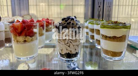 Desserti stratificati assortiti in bicchieri di vetro con fragole, panna montata, pezzi di biscotto scuro e guarnizioni verdi in una vetrina refrigerata. Foto Stock