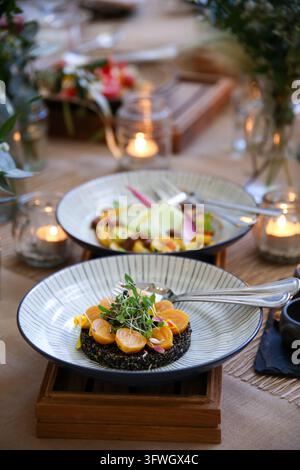 Due piatti gourmet elegantemente impiattati con microverdi e fette di agrumi sono posati su un tavolo di legno rustico adornato da candele e composizioni floreali. Foto Stock