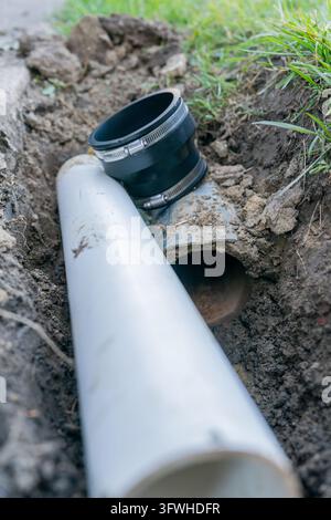 Un tubo esposto giace nella terra scavata. Un raccordo nero poggia accanto al tubo bianco. La terra circonda entrambi i tubi. L'erba verde incornicia la scena. Ideale per la costruzione Foto Stock