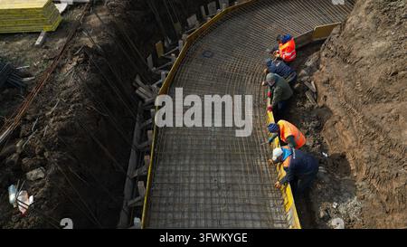 Grocka, Serbia – 20 ottobre 2026: Vista aerea di operai edili che installano reti di rinforzo in acciaio per una struttura in cemento curva in una trincea. Foto Stock