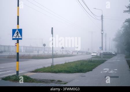 Otwock, Polonia – 16 ottobre 2026: Una fitta nebbia mattutina inghiotte una strada tranquilla e un percorso per cicli, costringendo i conducenti a rallentare e accendere le luci Foto Stock