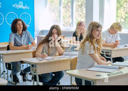 Gruppo di studenti delle superiori seduti a banchi in aula con muro blu che presenta formule matematiche. Gli studenti sono concentrati sullo studio, scriva Foto Stock