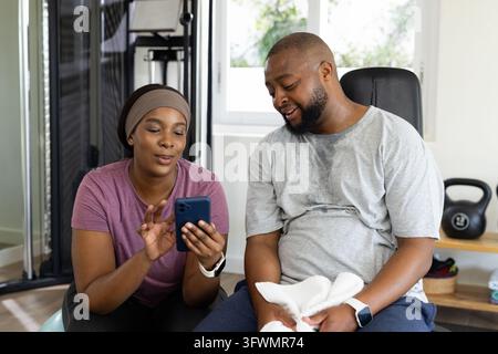 Coppia africana in abbigliamento sportivo seduta in una palestra casalinga, donna che mostra un telefono blu mentre l'uomo è appoggiato Foto Stock