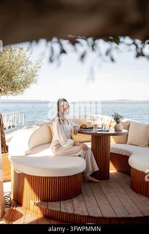 Donna d'affari sorridente con occhiali seduta accanto a un laptop aperto in un caffè sul mare. Un dolce, una bevanda fredda e la vista sul mare completano la rilassata scena all'aperto Foto Stock