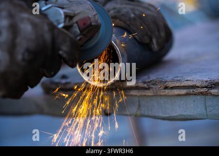 L'operaio dirige un disco abrasivo rotante contro l'estremità di un tubo d'acciaio su un banco da lavoro con scintille che cadono a cascata, illustrando la preparazione manuale dei tubi in me Foto Stock