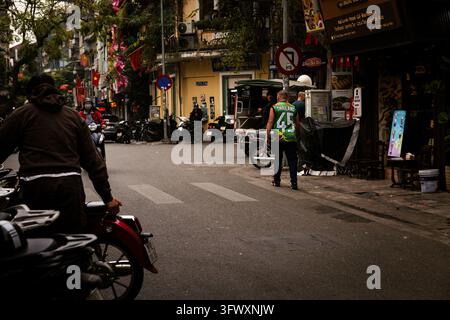 Hanoi, Vietnam - 16 gennaio 2026: Una scena di strada che mostra pedoni e motociclette vicino a una farmacia e negozi con vari cartelli e bandiere. Foto Stock