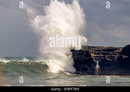 Un enorme spruzzo di acqua marina bianca e schiuma erompe nell'aria mentre un'onda forte dell'oceano si schianta direttamente contro una scogliera rocciosa scura sulla costa. Foto Stock