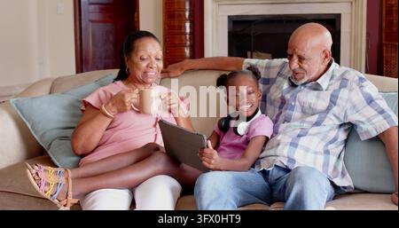 Coppia di anziani afroamericani con nipote seduti sul divano in salotto mentre usano un tablet. Famiglia, anziano, bambino, relax, accogliente, moderno, interg Foto Stock