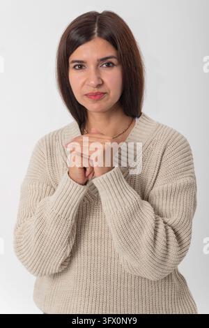 Donna in piedi su sfondo bianco con maglione di lana beige a scollo a V, mani giunte, espressione pensierosa e concentrata, ritratto di studio di una penne Foto Stock