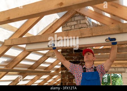 Costruttore che utilizza un livella a bolla per verificare l'allineamento orizzontale delle nuove travi del tetto in legno durante la costruzione residenziale, garantendo una misurazione accurata Foto Stock