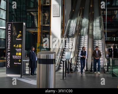 Una guardia di sicurezza è di guardia accanto alle scale mobili mentre due impiegati di ufficio in camicia bianca scendono verso l'atrio principale della reception all'interno del concorso pubblico all'aperto del Leadenhall Building (conosciuto informalmente come The Cheesegrater) nella City di Londra, Regno Unito, il 18 settembre 2026 Foto Stock