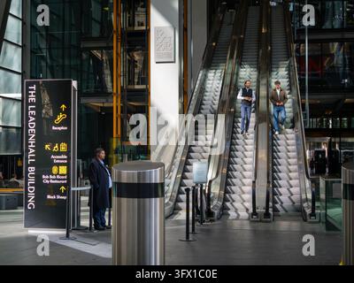 Una guardia di sicurezza è di guardia accanto alle scale mobili mentre due impiegati di ufficio in camicia bianca scendono verso l'atrio principale della reception all'interno del concorso pubblico all'aperto del Leadenhall Building (conosciuto informalmente come The Cheesegrater) nella City di Londra, Regno Unito, il 18 settembre 2026 Foto Stock