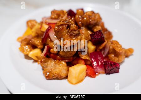 Maiale dolce e aspro con ananas, frutto del drago, cipolla rossa e peperoncino su un piatto di ceramica bianca, piatto classico in stile cantonese servito in un ambiente ristorante moderno Foto Stock