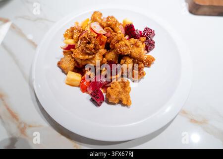 Pollo croccante dolce e piccante con ananas a cubetti, frutto del drago rosso e cipolla rossa affettata, servito su un piatto di ceramica bianca sopra un tavolo di marmo bianco in un ambiente moderno di ristorante Foto Stock