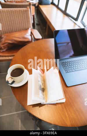 Quaderno aperto con pagine scritte a mano e una tazza di ceramica con caffè nero su un tavolo rotondo di legno accanto a un laptop argentato in un caffè moderno con sedie di vimini, grandi finestre e luce naturale del giorno che illumina l'accogliente ambiente di lavoro Foto Stock