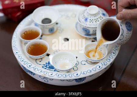 Una mano versa un tè oolong color ambra da una teiera di porcellana blu e bianca in una piccola tazza di ceramica su un vassoio da tè tradizionale cinese, circondato da tazze assortite, una gaiwan con coperchio e un foro di drenaggio, evocando il rituale sereno della preparazione del tè Gongfu in un ambiente domestico. Foto Stock