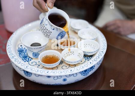 Una mano versa un tè oolong ambrato scuro da una delicata teiera di porcellana blu e bianca traforata in piccole tazze da degustazione abbinate disposte su un vassoio circolare tradizionale, mostrando l'estetica raffinata e il rituale della cerimonia del tè Gongfu cinese in un ambiente interno caldo. Foto Stock