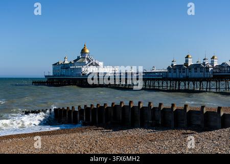 EASTBOURNE PIER E ACQUE COSTIERE DAL PEBBLE BEACH, REPORTAGE DI VIAGGIO, EASTBOURNE, INGHILTERRA: L'intera lunghezza del padiglione storico del molo e del passaggio si estende in acque marine agitate oltre una groyne di legno sotto il sole splendente. INFO: Eastbourne è una raffinata città termale sulla costa sud-orientale dell'Inghilterra, famosa per le sue imponenti scogliere di gesso. Con un classico molo vittoriano, giardini lussureggianti sul lungomare e una luce solare eccezionale, funge da drammatico ingresso a Beachy Head. Foto Stock